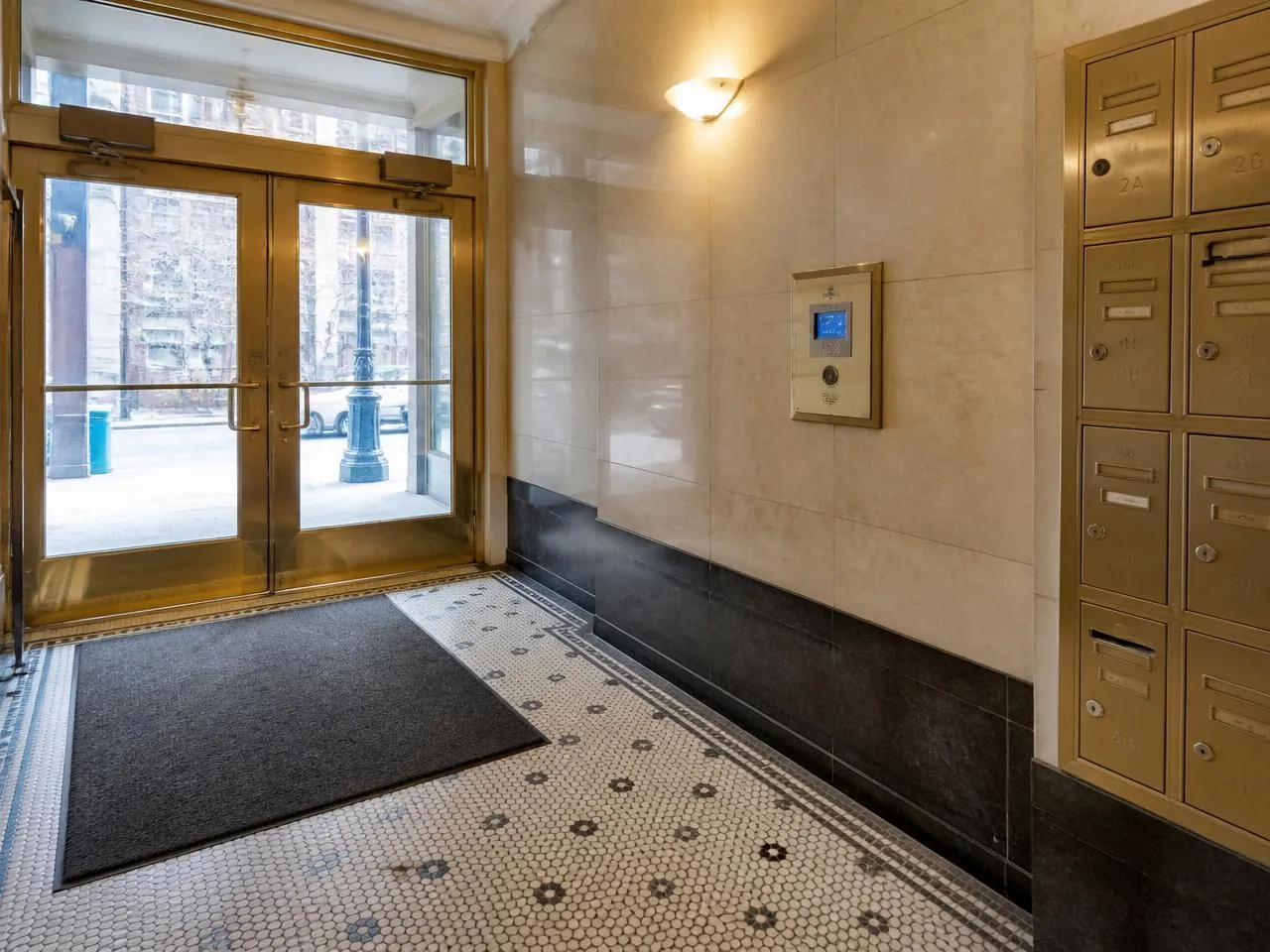 Renovated New York City apartment building vestibule with refinished brass entry doors, mosaic tile floor, and flush intercom panel