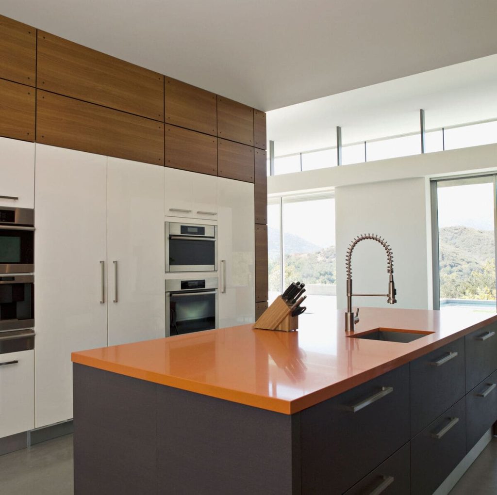 Modern kitchen with white cabinets, built-in stainless steel appliances, orange countertop island, sink with gooseneck faucet, and knife block, with large windows in the background.