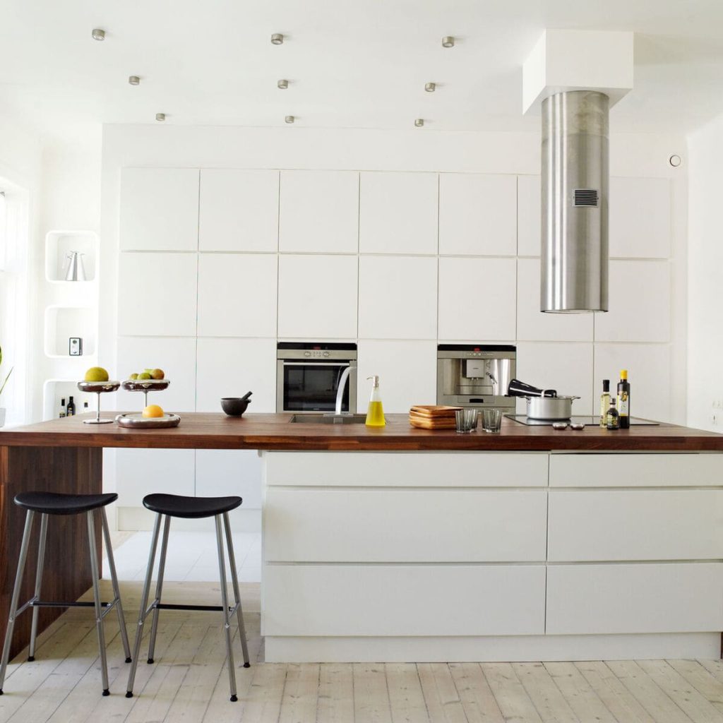 Modern white kitchen with minimalist cabinets, a wooden countertop island, two black stools, and built-in stainless steel appliances. Fruit bowls and kitchenware are on the counter.